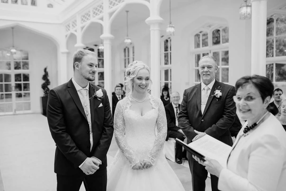 A wedding ceremony with a bride and groom standing with an officiant, facing each other, inside a bright, elegant venue with large windows and white decor, surrounded by seated guests.