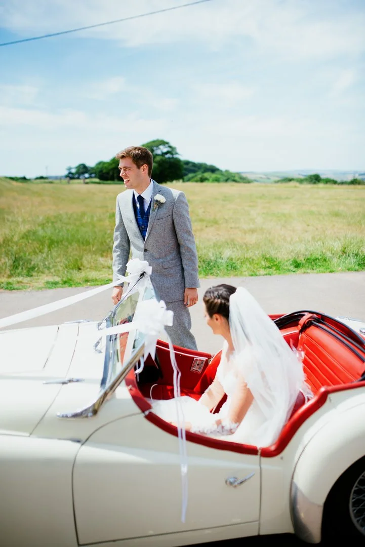 A bride sitting in a vintage white convertible car with red interior, while a groom stands outside in a gray suit, smiling, in an open field under a partly cloudy sky.