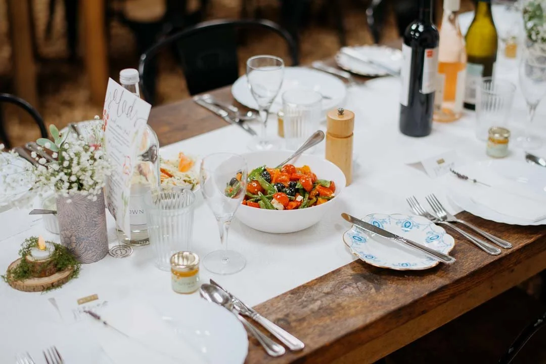 A rustic wooden dining table set for a meal with white plates, silverware, glasses, and floral centerpieces. There is a bowl of mixed vegetables, bottles of wine, and small jars on the table.