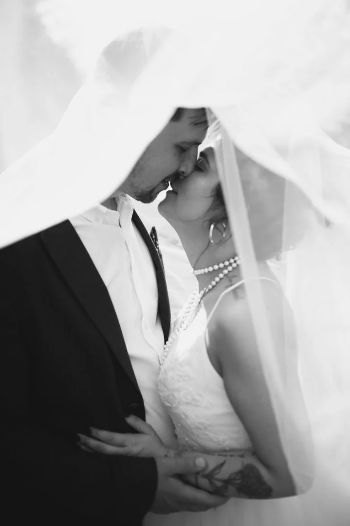 A black-and-white photograph of a bride and groom under a wedding veil, touching noses with their eyes closed, sharing a tender moment during their wedding.