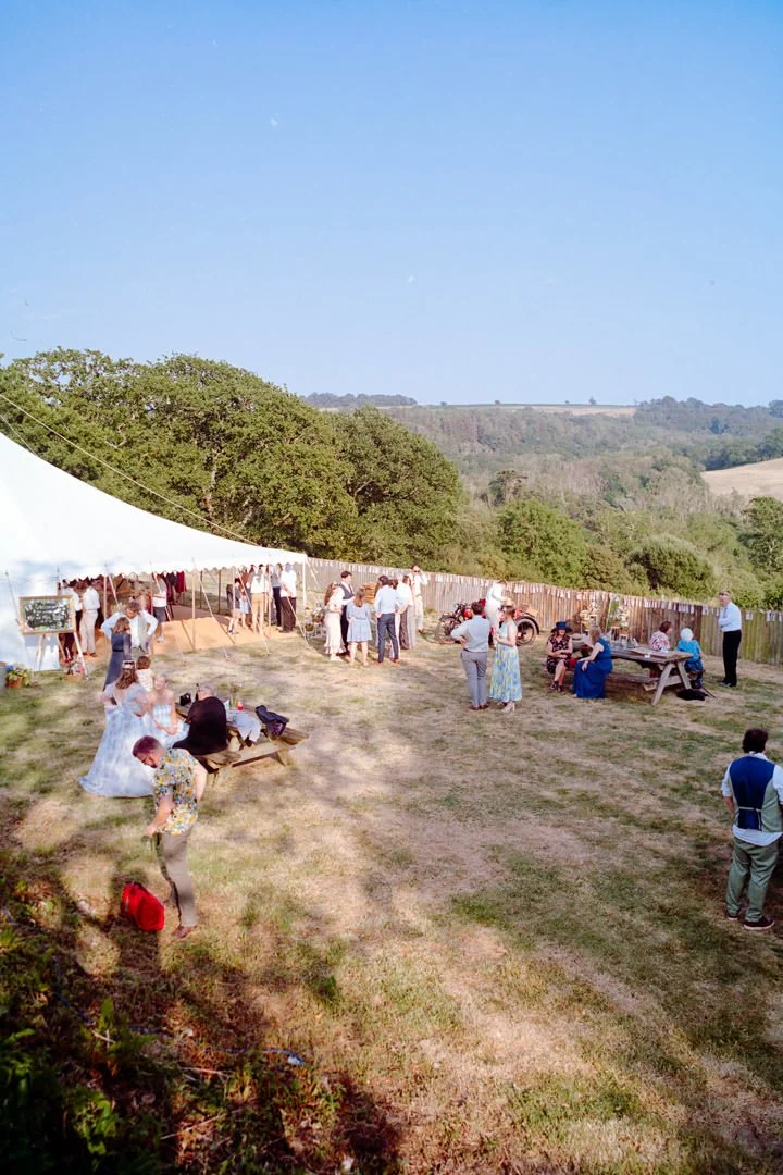 People gathered outdoors near a large white event tent on a sunny day, with some seated at picnic tables and others standing and mingling in a grassy area.