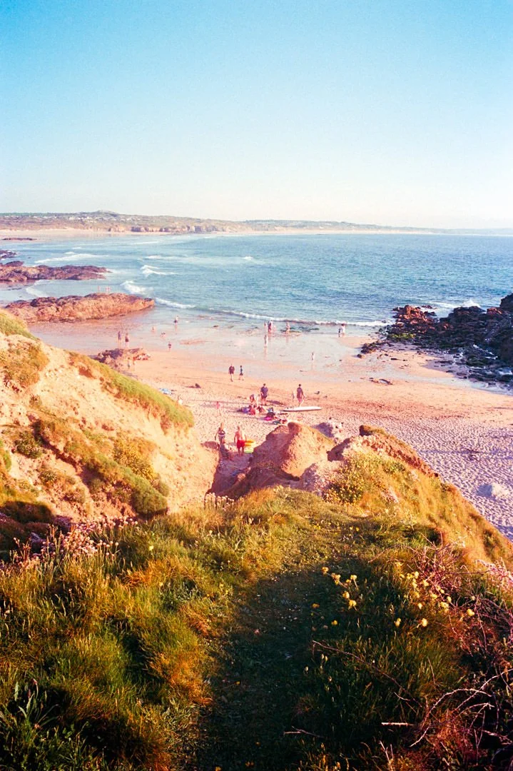 A scenic view of a sandy beach with rocks, greenery, and cliffs, with people swimming and relaxing in the ocean and on the shoreline under a clear blue sky.