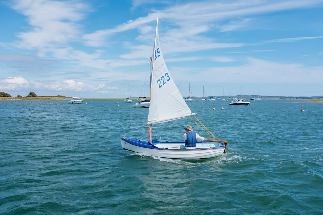 A person sailing a small sailboat on a calm body of water with other boats in the background under a partly cloudy sky.