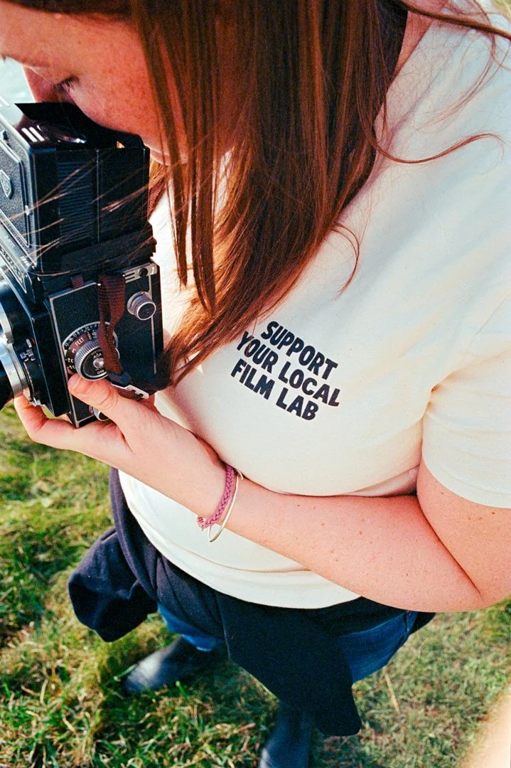Person with long brown hair wearing a beige T-shirt with the words 'Support Your Local Film Lab' and holding a vintage camera outdoors on grass.