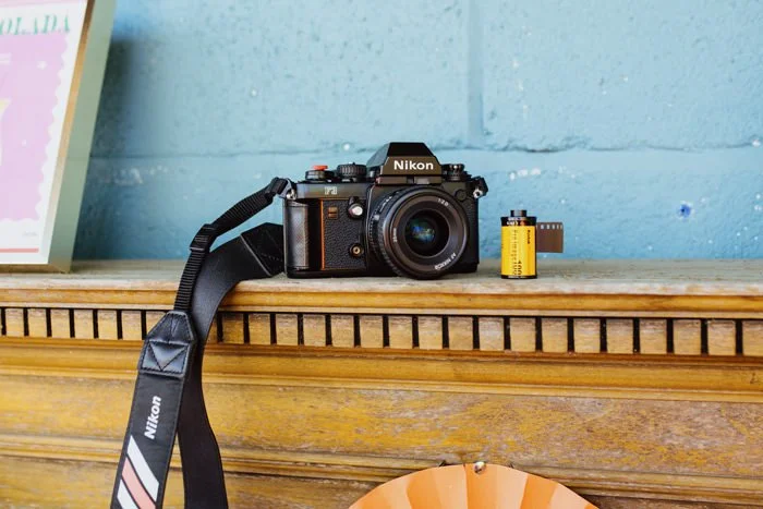 A Nikon film camera with a strap, a roll of film, and a small yellow container on a wooden surface against a blue wall.