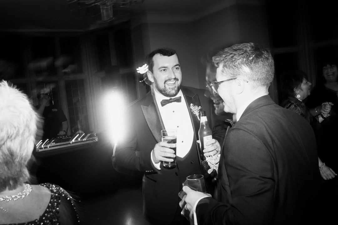 People in formal attire socializing at a party, with a man in a tuxedo holding a drink and smiling.