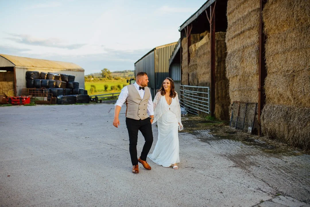 A bride and groom walking hand in hand on a farm, with hay bales and farm buildings in the background.