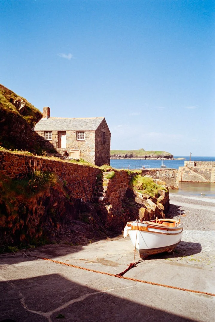 A small white boat pulled up on a concrete slipway at a harbor, with a stone house on a hill above and the ocean in the background.