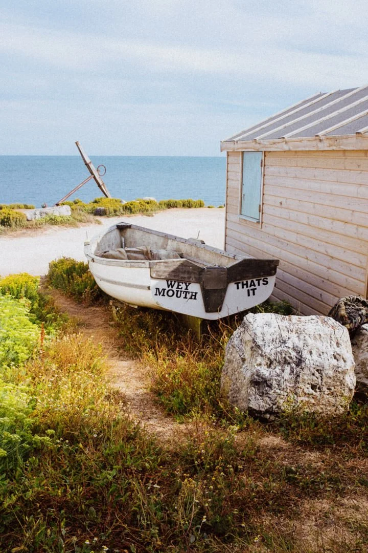 A small boat on a beach with a sign that reads "WEY MOUTHTHATS IT." The boat is resting on the ground near lush greenery and a large rock. In the background, there is a body of water, a lamppost with a tilted light, and a wooden building.