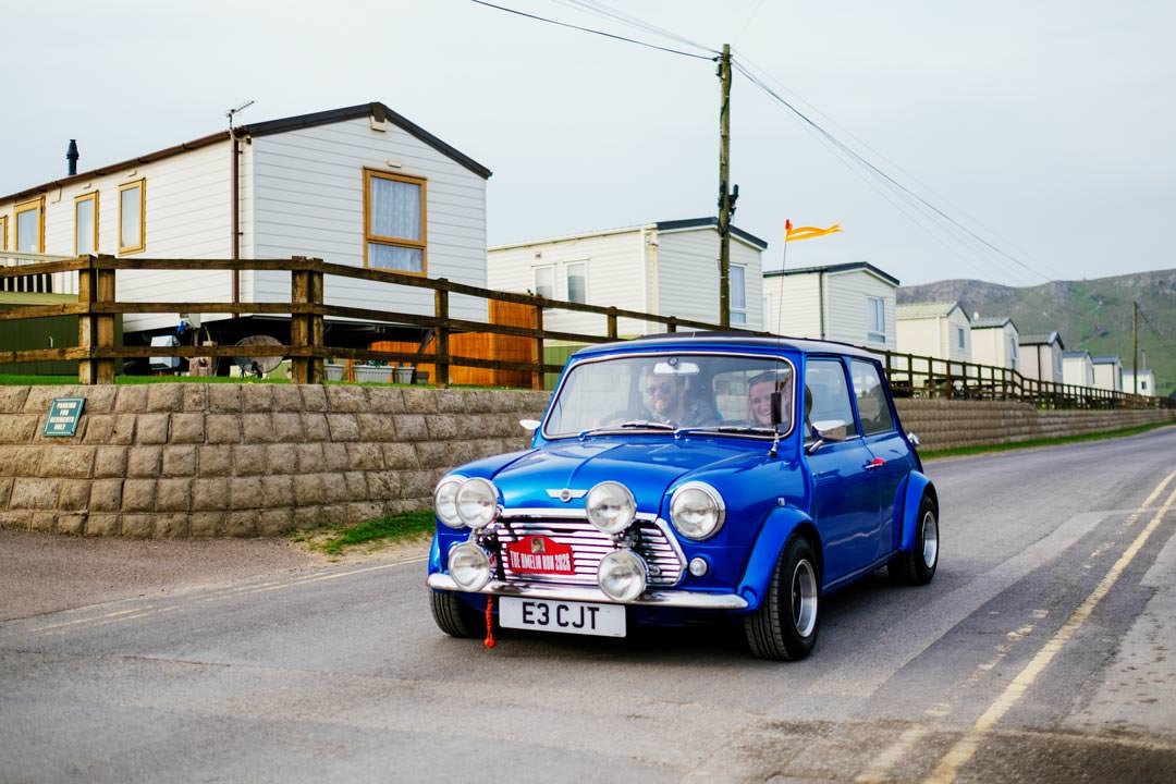 A blue classic Mini Cooper with multiple auxiliary lights on the front, driving along a rural road with mobile homes and green mountains in the background.