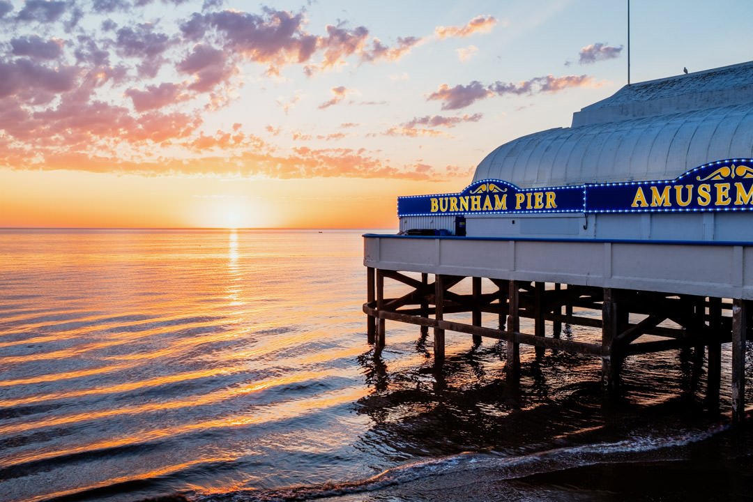 Sunset over the water at Burnham Pier amusement park, with clouds in the sky and the pier's building partially visible on the right.