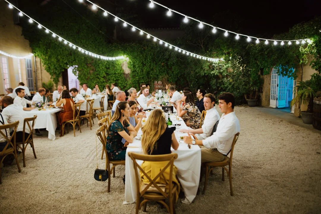 A large outdoor dinner party at night with numerous guests seated at long tables decorated with white tablecloths, candles, and flowers, illuminated by string lights overhead, with lush green foliage and a blue door in the background.