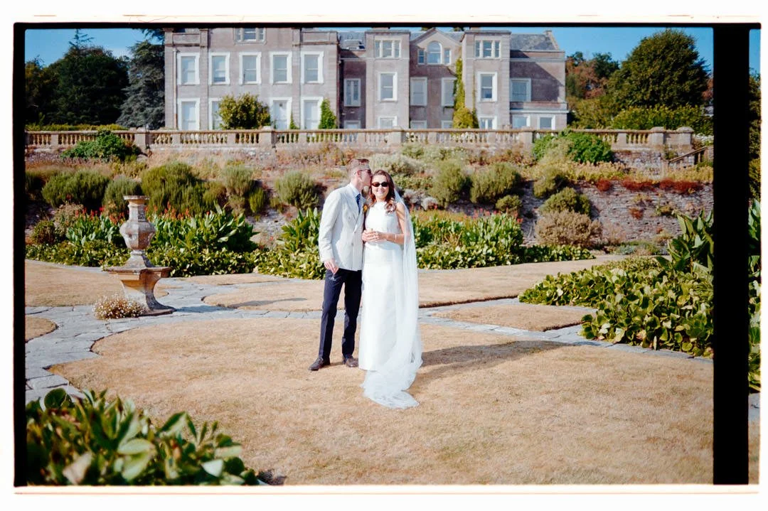 A bride and groom at a wedding venue outdoors, with the bride wearing a white gown and sunglasses, and the groom dressed in a light-colored suit. They are standing on a landscaped garden with a large house in the background, under clear blue skies.
