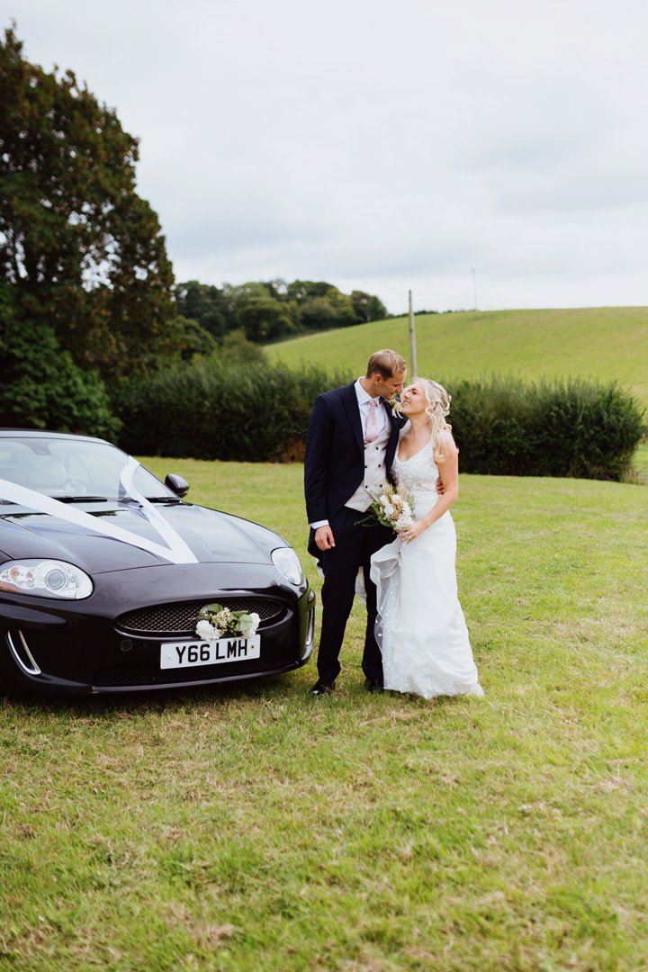 A bride and groom in wedding attire standing on a grassy field next to a decorated black car with white ribbons and flowers, with green hills and trees in the background.