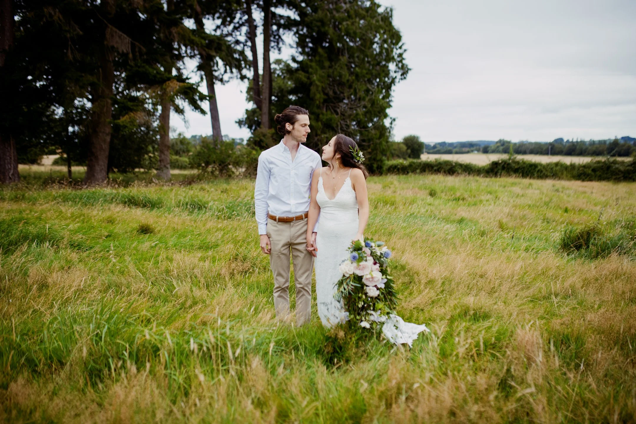 A bride and groom holding hands in a green field, with trees in the background, during a wedding photoshoot on an overcast day.