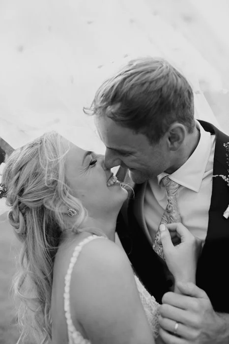 Black and white photo of a smiling bride and groom close together, touching noses, at their wedding.