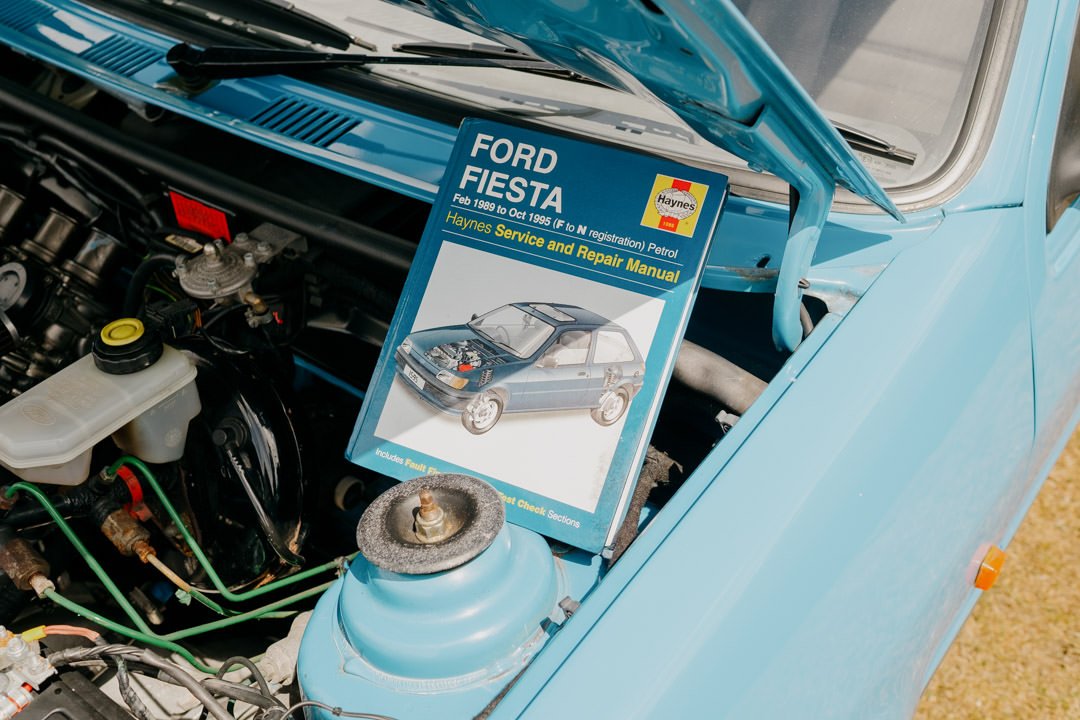 Open hood of a light blue Ford Fiesta showing engine components, with a manual titled 'Ford Fiesta Service and Repair Manual' resting on the engine bay.