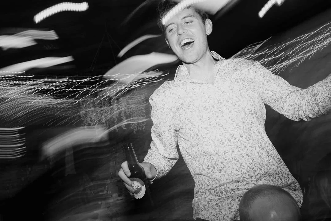 Black and white photo of a man celebrating at a party, holding a beer bottle, smiling, with blurred lights in the background.