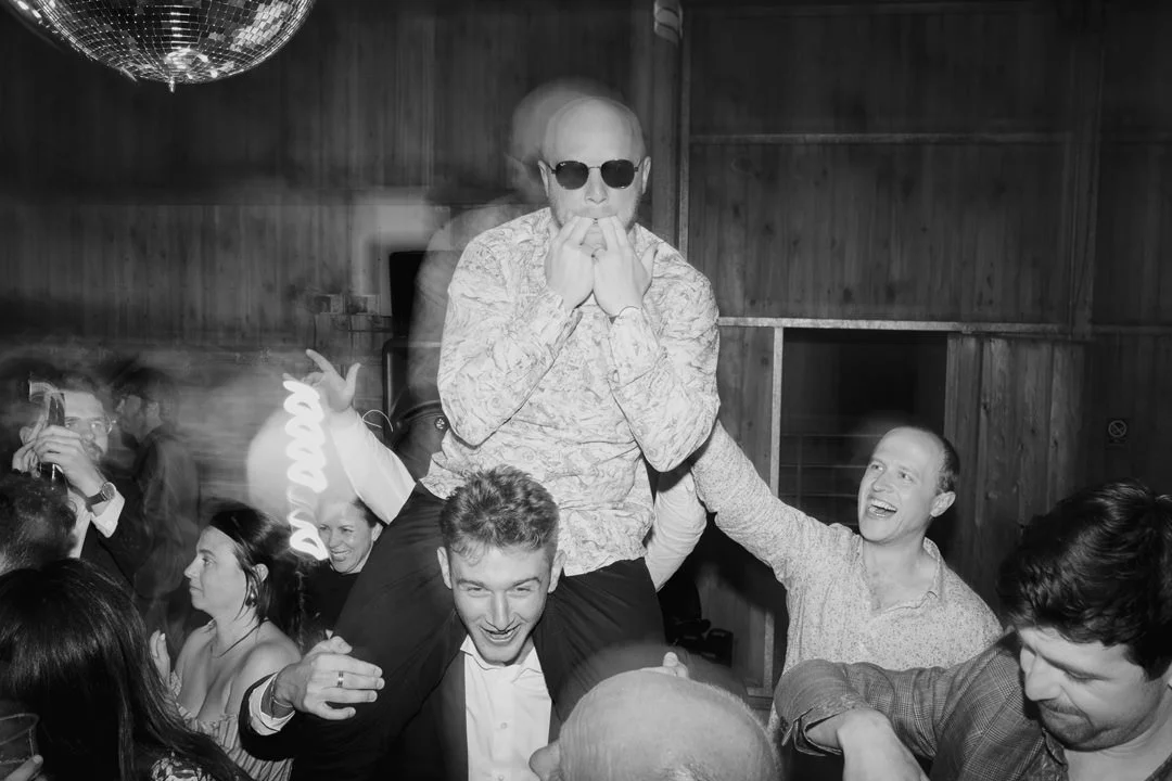 Group of people at a party, with one person wearing sunglasses sitting on someone's shoulders, others are smiling and interacting.