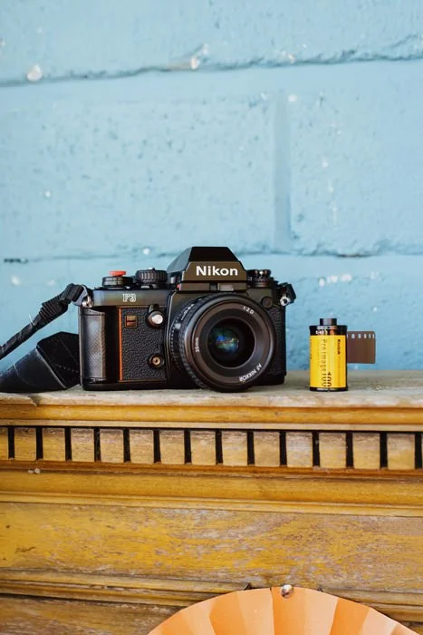 A Nikon film camera with a strap sits on a wooden surface next to a roll of film, against a light blue brick wall background.