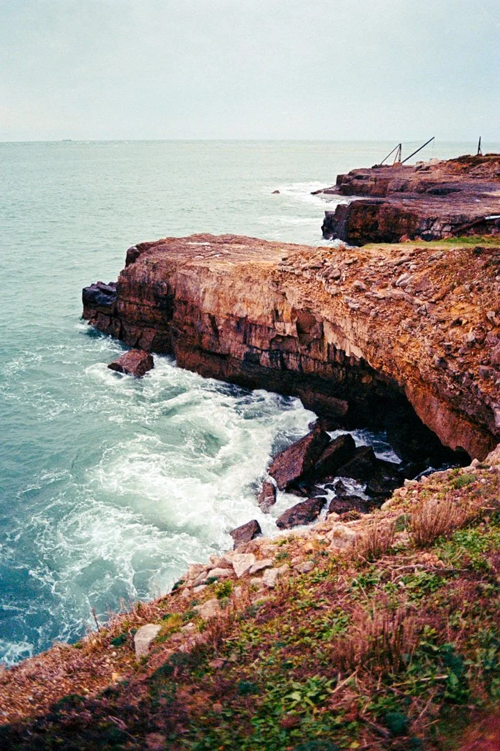 Rocky ocean shoreline with waves crashing against cliffs and sparse vegetation in the foreground.