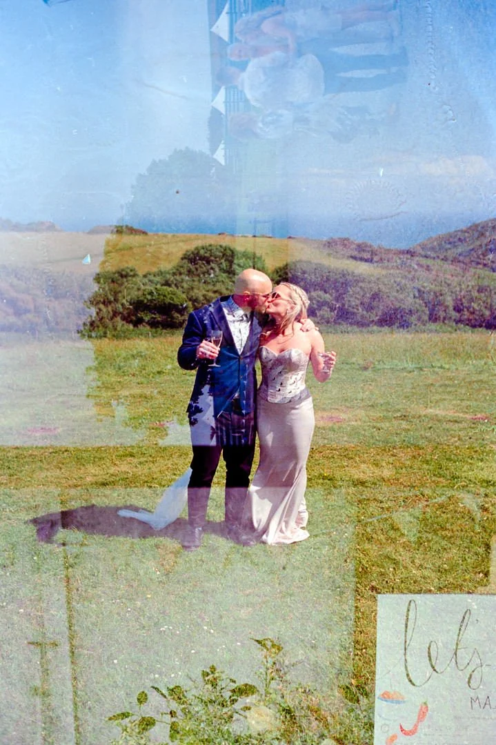 A couple dressed in formal attire sharing a kiss outdoors on a sunny day, holding glasses of wine, with a grassy field and hills in the background.
