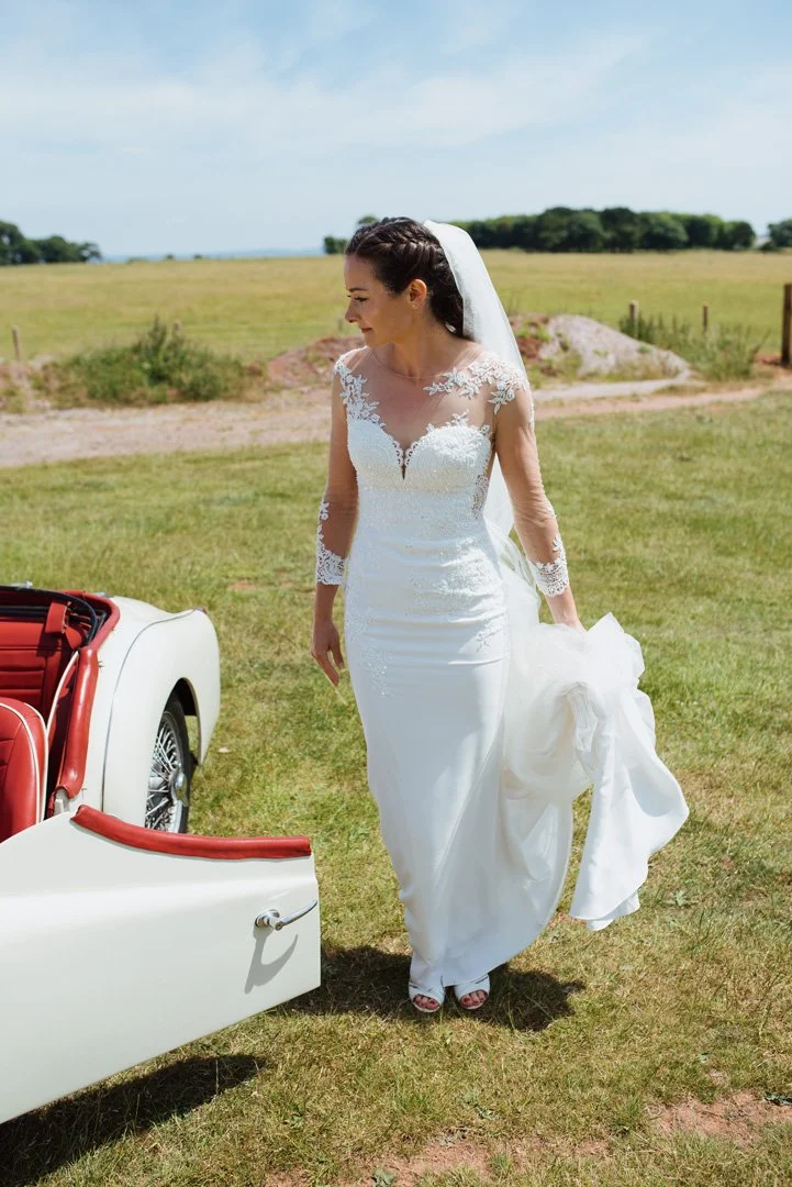 A bride in a white wedding dress with lace details and a veil stands outdoors on grass, holding her gown, beside a vintage white car with red interior, in a rural setting under a blue sky.