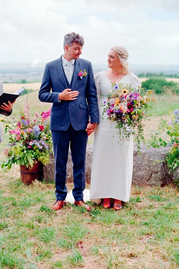 A couple in wedding attire during their outdoor wedding ceremony, holding hands and standing beside a flower arrangement, with an officiant holding a book in the background.