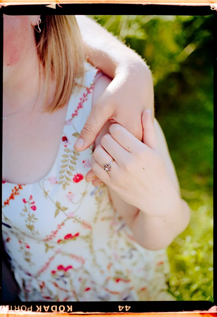Close-up of a woman with blonde hair wearing a floral dress, showing her hand with a ring on her finger and adjusting her shoulder, outdoors with greenery in the background.