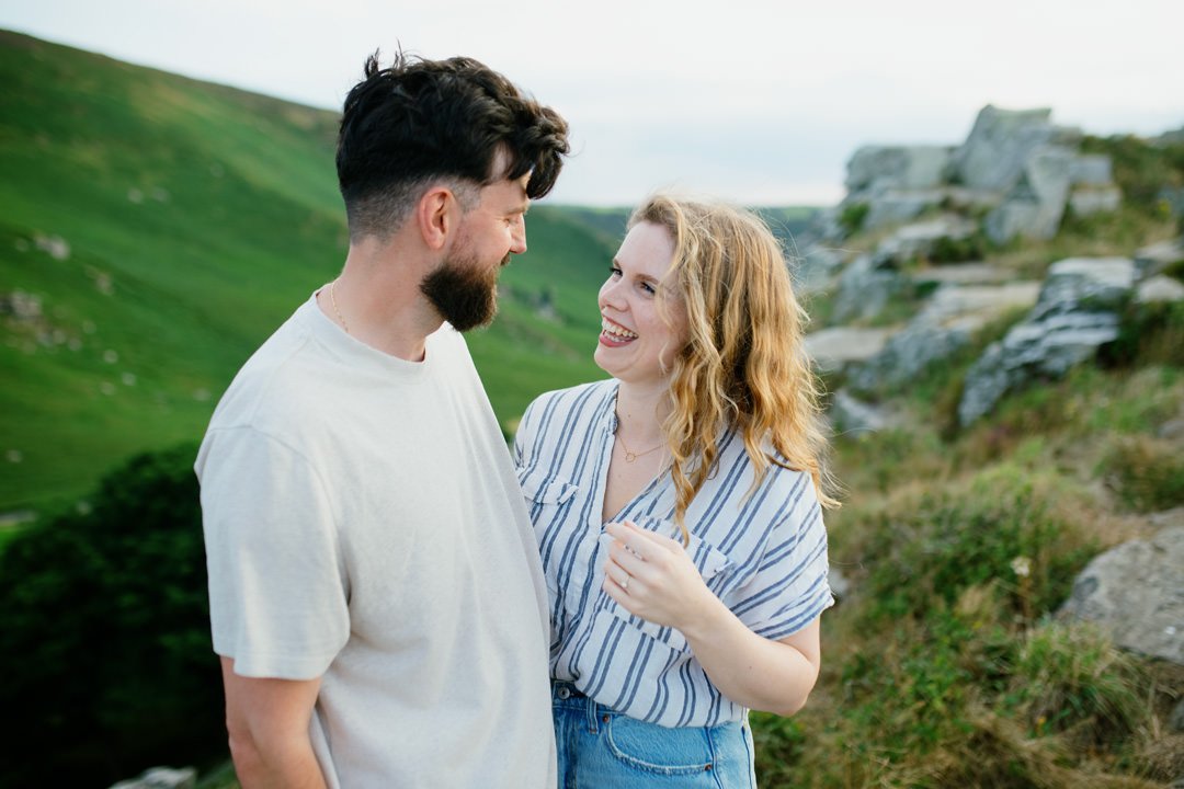 A happy couple outdoors in a grassy, hilly area with rocks, smiling and looking at each other.