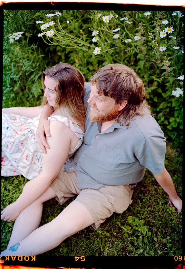 A man and woman sitting on grass surrounded by green foliage and white daisies, sharing a moment of closeness and smiles.