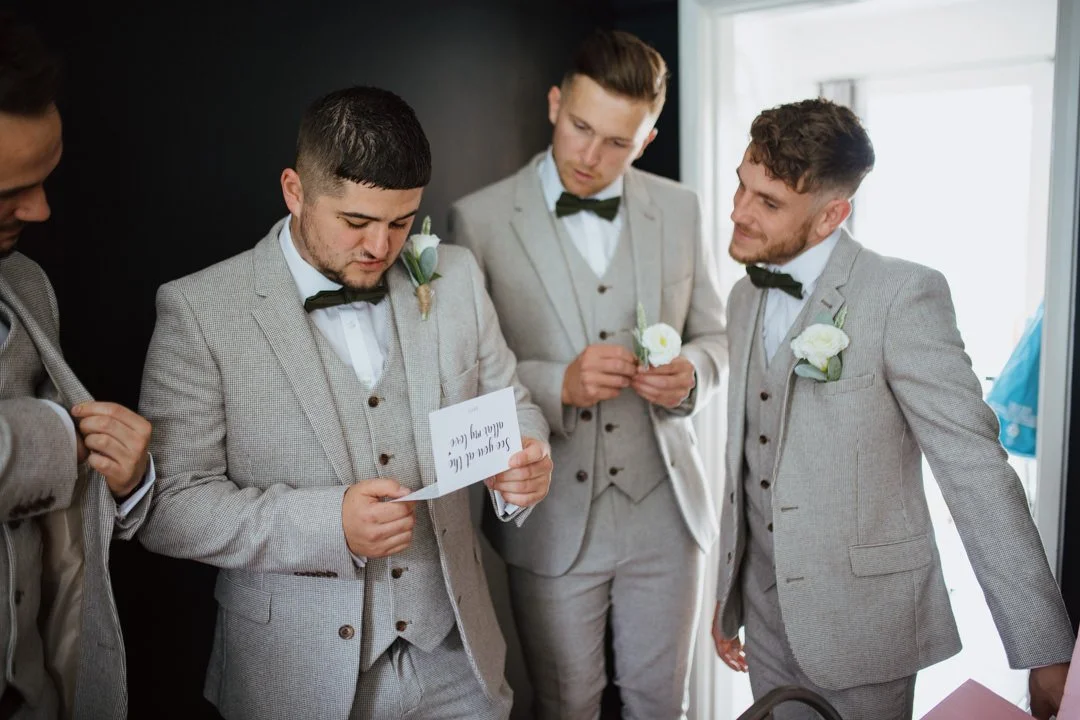 groomsmen waring grey suits reding a letter together in a large room with a wooden floor