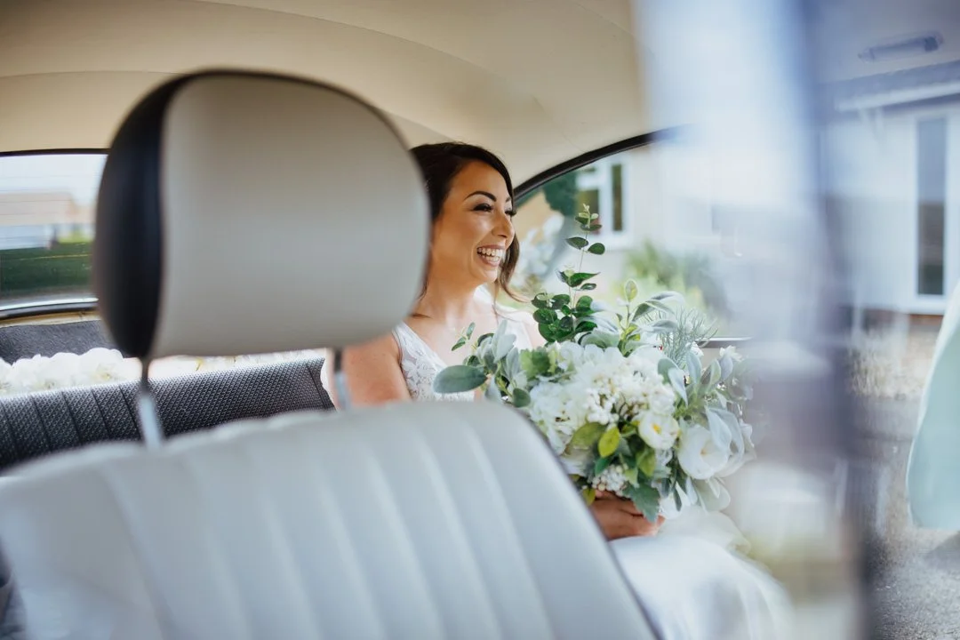bride holding large bunch of flowers sat on the back seat of a car. she is smiling looking at her bridesmaids