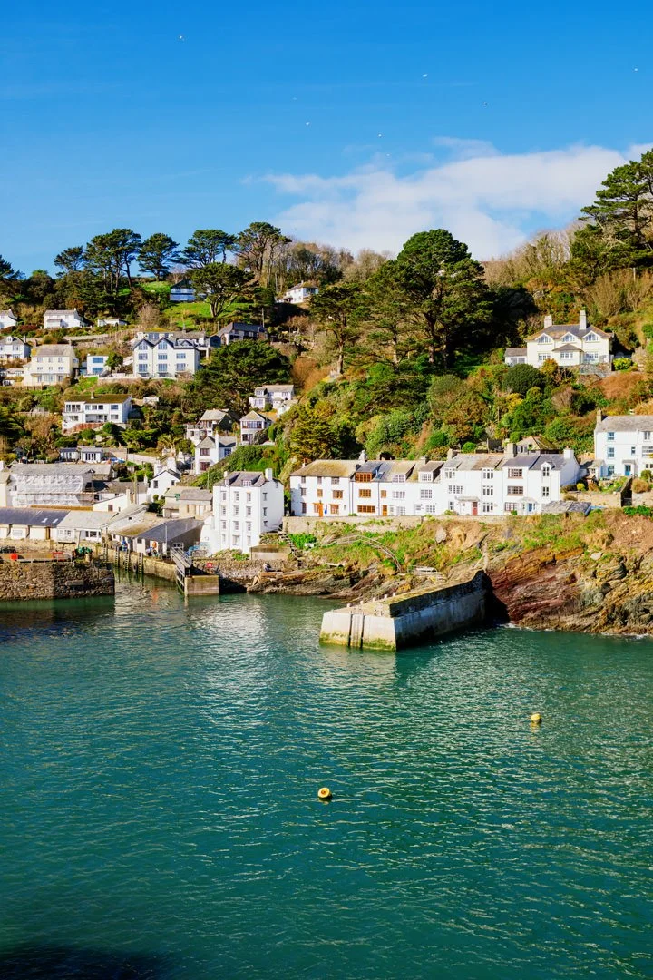 Coastal scene with a hillside filled with white and pastel-colored houses overlooking the ocean, blue sky with a few clouds, and calm water in the foreground.