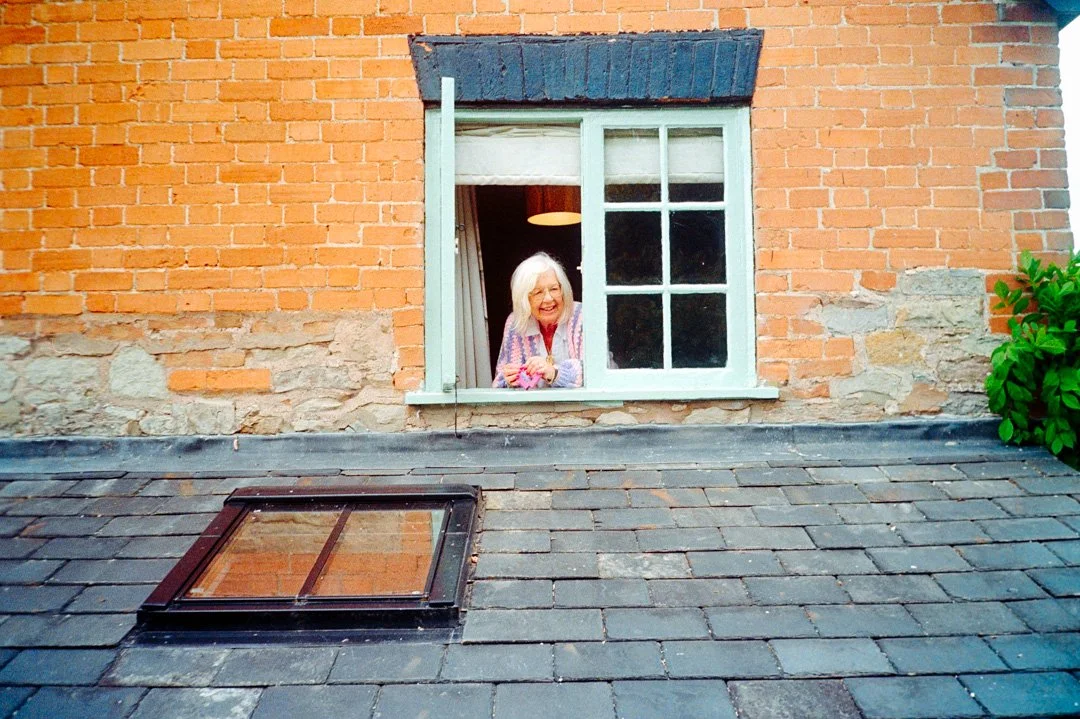 An elderly woman smiling and looking out of a window of a brick house.
