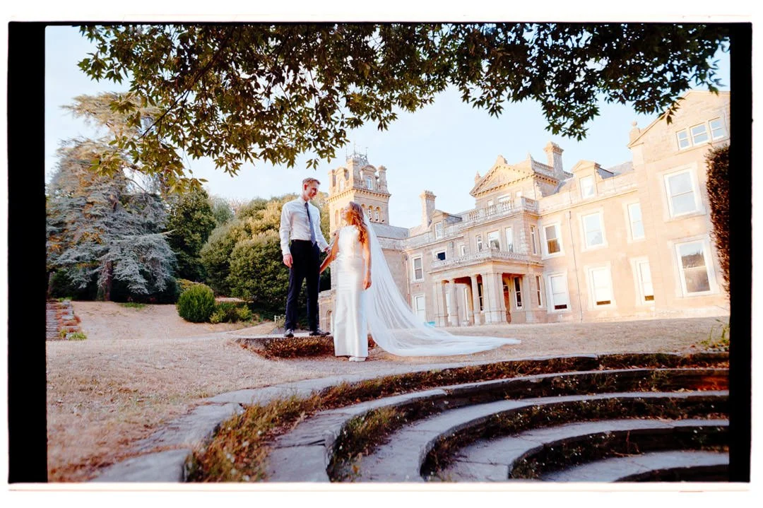 A bride and groom holding hands outdoors in front of a large historic mansion surrounded by trees.