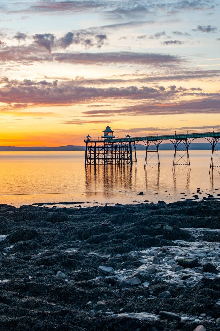 Sunset over a bay with a pier and pavilion, rocky shoreline in the foreground, colorful sky with scattered clouds.