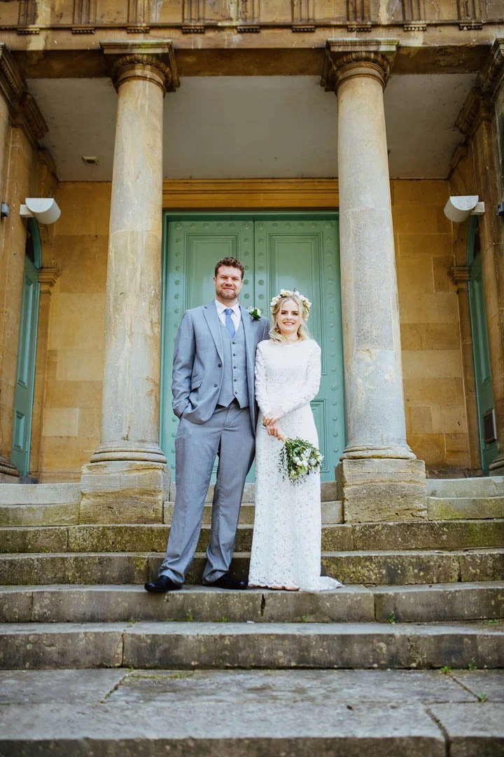 A bride and groom standing on steps in front of a historical church building with large wooden doors and two stone columns. The bride is holding a bouquet and wearing a lace wedding dress and floral crown. The groom is dressed in a gray suit with a b