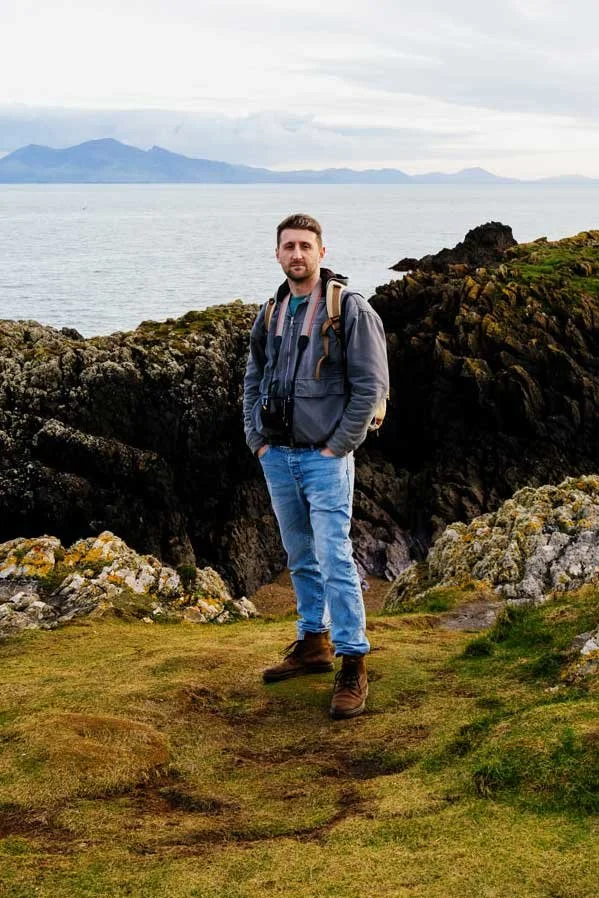 A man standing on grassy terrain with rocky formations, near a body of water, mountains in the background, overcast sky, wearing a gray jacket, blue jeans, brown hiking boots, and carrying a backpack.