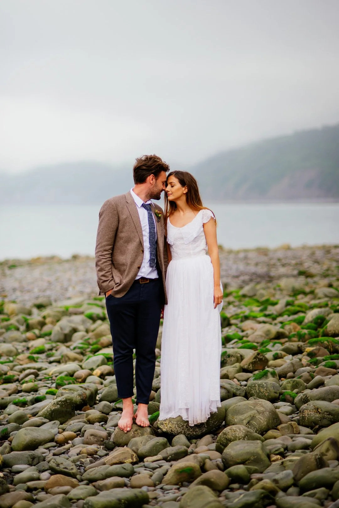 bride and groom stood on stoney beach with large cliff behind them under grey cloudy sky