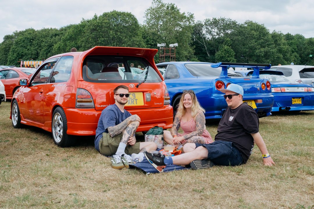 Three people sitting on a picnic blanket at a car show, with two modified sports cars behind them, on a grassy field with trees in the background.