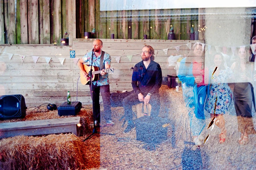 A guitarist and a seated man performing at an outdoor event with a wooden fence and bunting in the background.