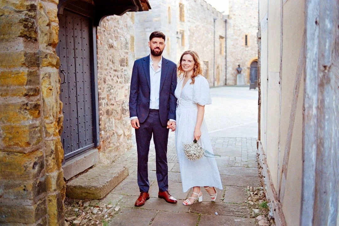 A man and woman standing hand-in-hand outdoors near a stone building, smiling at the camera. The man is wearing a dark suit, white shirt, and brown shoes. The woman is in a white dress, holding a small bouquet of flowers, and wearing white heels.