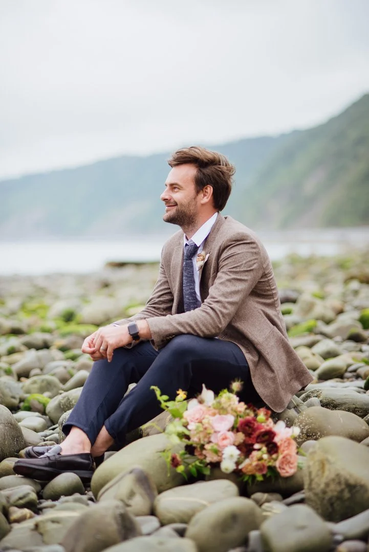 wedding groom sat on large rock with flowers next to his feet