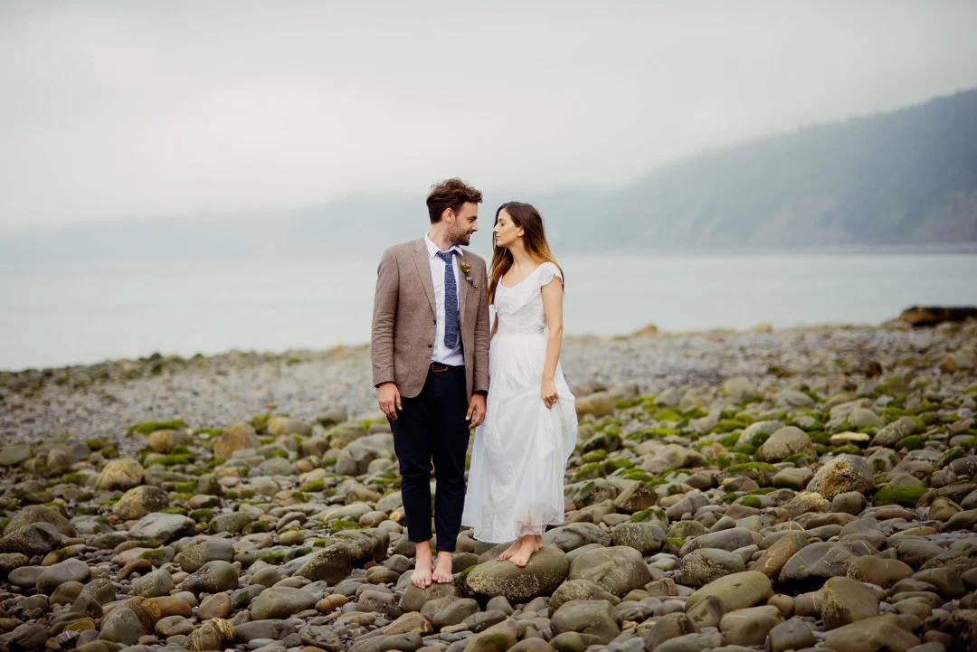 A bride and groom walking barefoot on a rocky beach, looking at each other.