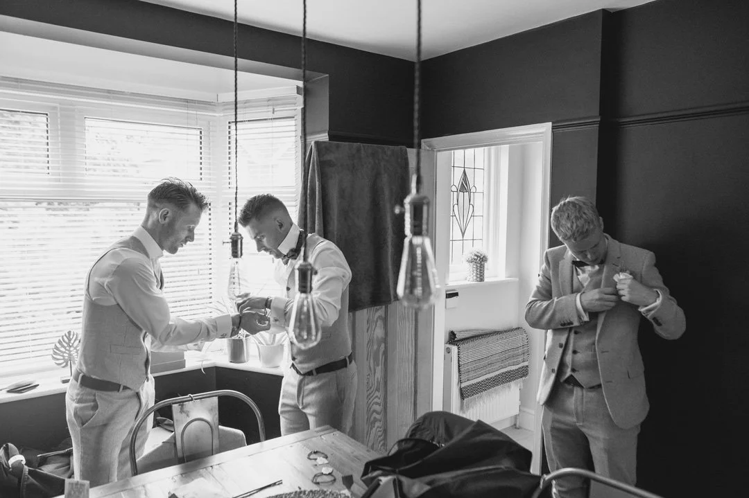 three groomsmen putting on ties in large room with wooden floor. There are industrial lights hanging from the celling