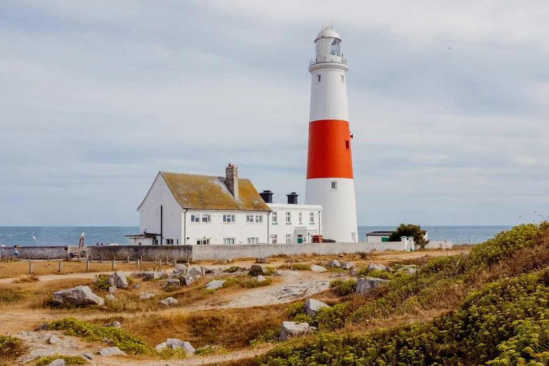 A lighthouse with a red and white striped tower next to a white house, situated near the coast with an ocean in the background and rocky, grassy land in the foreground.