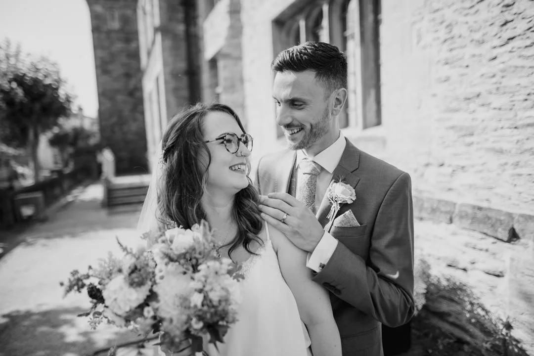 black and white photo of bride and groom, they stood outside large stone building smiling
