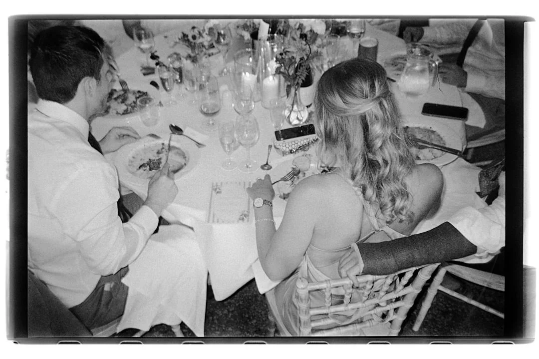 People sitting around a dinner table with wine glasses, flowers, and food, engaged in a meal, in black and white.