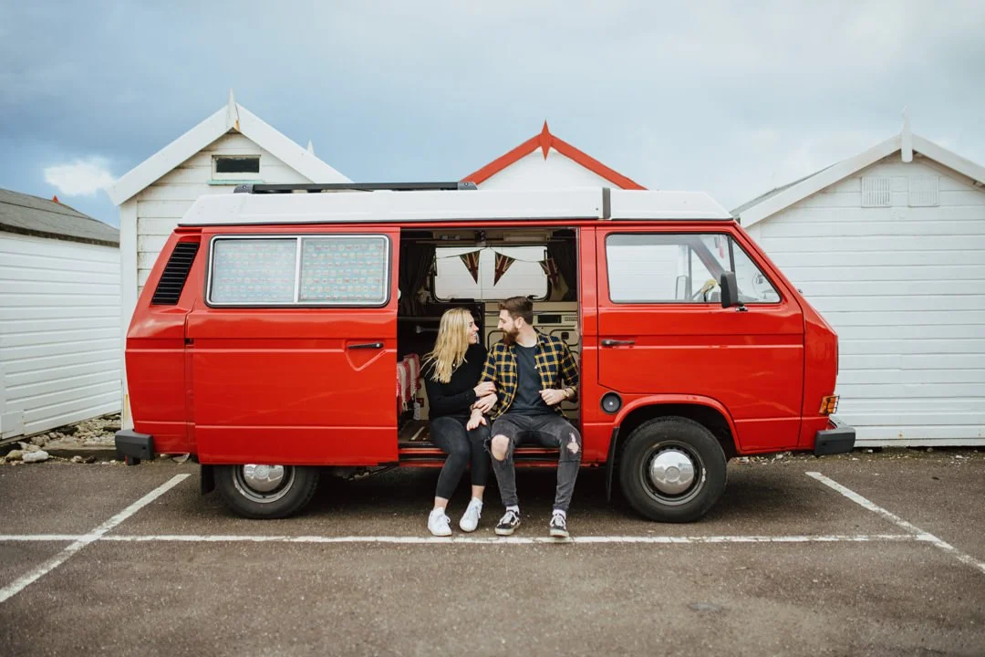 A couple sitting and talking in front of a red camper van parked in a lot with white beach huts in the background.
